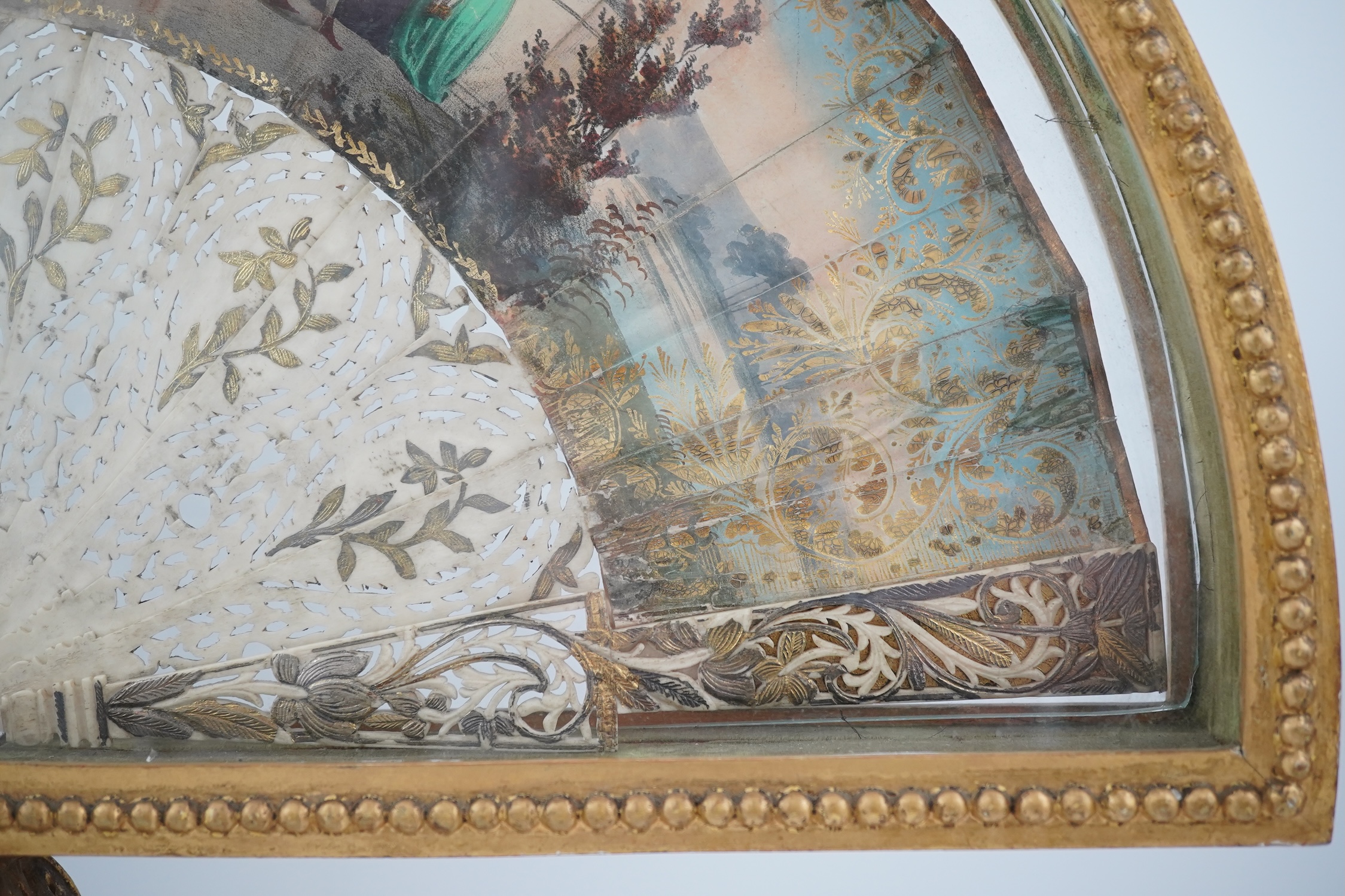 Two giltwood fans and display cases, one on a pedestal base housing an 18th century French figural hand painted fan with bone and gilt decorated guards and sticks, the other set with paste gilt metal guards on mother of